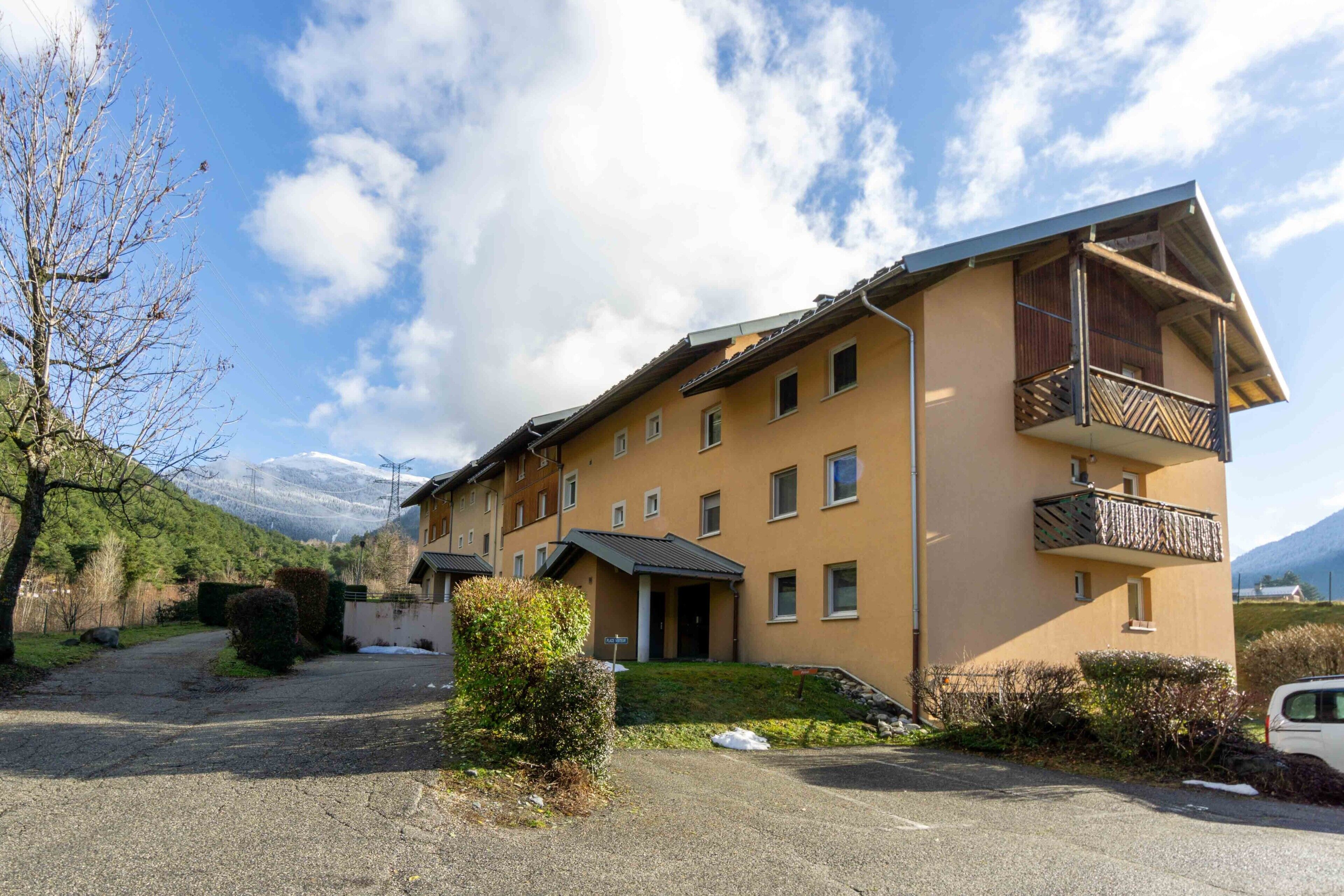 A multi-story building with a balcony, a driveway, and a mountain in the background.