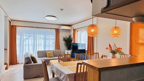 Una sala moderna con un sofá, una mesa de comedor y sillas. Hay un televisor sobre un mueble bajo, una planta en maceta y una ventana con cortinas.
