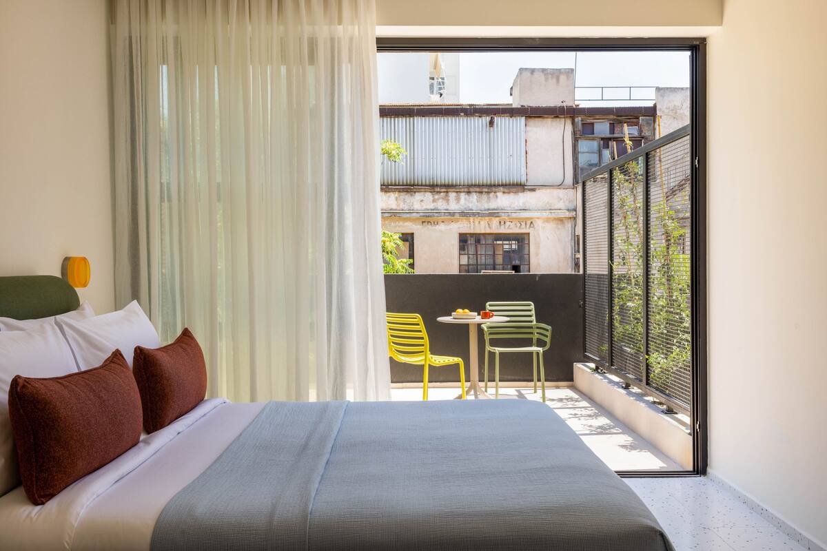 A bedroom with a bed, a window with sheer curtains, a balcony with a table and chairs, and a view of buildings outside.