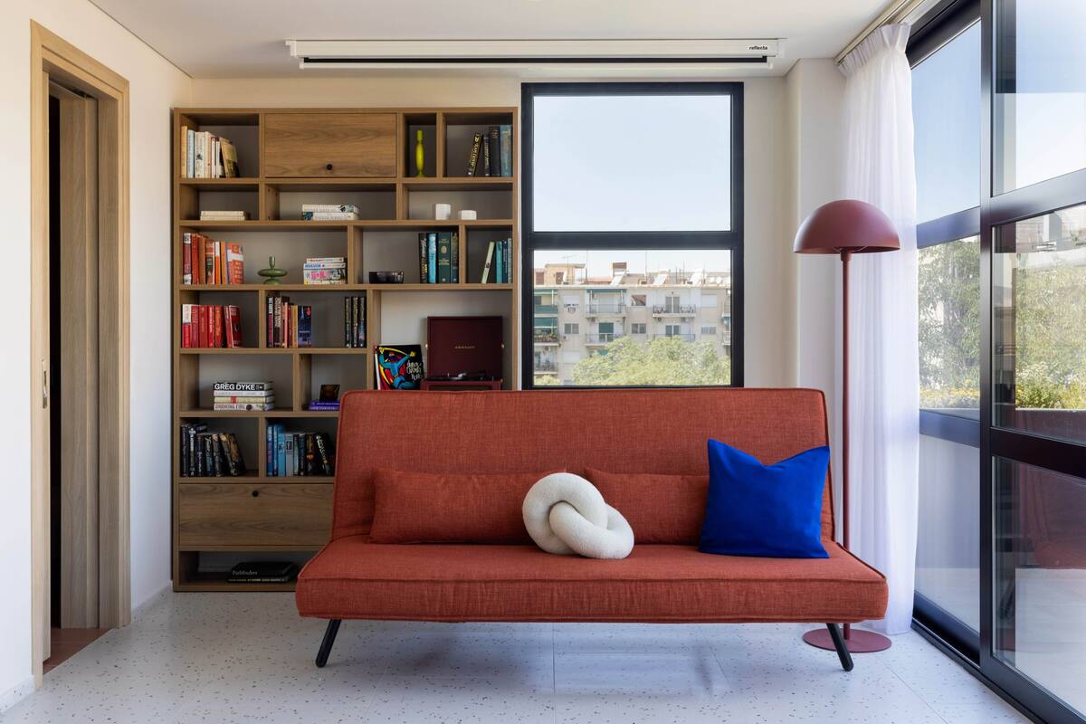 A modern living room with a red sofa, a wooden bookshelf, and a large window with a view of buildings.