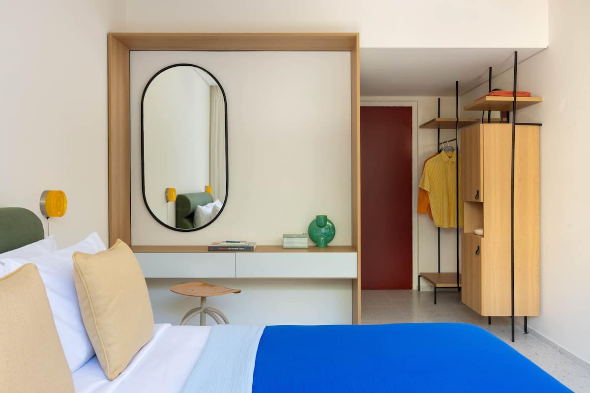 A hotel room with a bed, a bedside table, a mirror, a wardrobe, and a small stool.