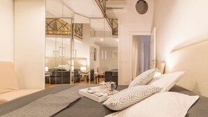 A modern bedroom with a large bed, a bedside table with a tray of breakfast items, and a view into the living area through a glass partition.