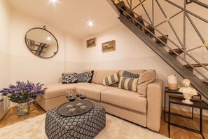 A living room with a beige sofa, patterned ottoman, and a round mirror. A staircase with a metal railing is visible in the background. - Campo de’ Fiori 35 - modern and quiet, in the center of Rome (Roma)