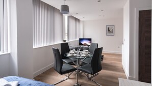 Un espace repas moderne avec une table en verre et des chaises noires, un téléviseur en arrière-plan et une grande fenêtre avec des stores.