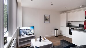 Un salon moderne avec un téléviseur à écran plat, une table basse blanche et un canapé gris. On aperçoit, au fond, une cuisine avec des meubles blancs et une poubelle en acier inoxydable.