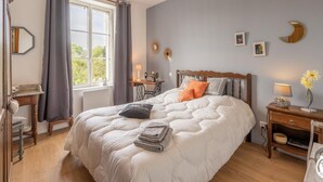 Een slaapkamer met een groot bed, een houten nachtkastje, een bureau en een stoel. Er is een raam met gordijnen, een hanglamp en decoraties aan de muur.