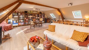Een gezellige zolderkamer met een hoge nok, houten balken, een grote boekenkast, een bank en een eettafel.
