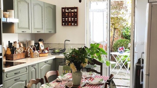 Een keuken met lichtgroene kastjes, een eettafel gedekt met een rood-wit geruit tafelkleed, en een vaas met bloemen.