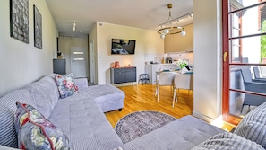 A modern living room with a sofa, a dining area with chairs, a kitchen with a TV, and a large window.