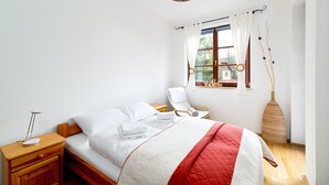 A neatly made bed with white linens and a red and white quilt, a wooden nightstand with a lamp, a wicker chair, and a window with curtains.