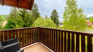 A balcony with a wooden railing, wicker furniture, and a view of green trees and houses in the background.