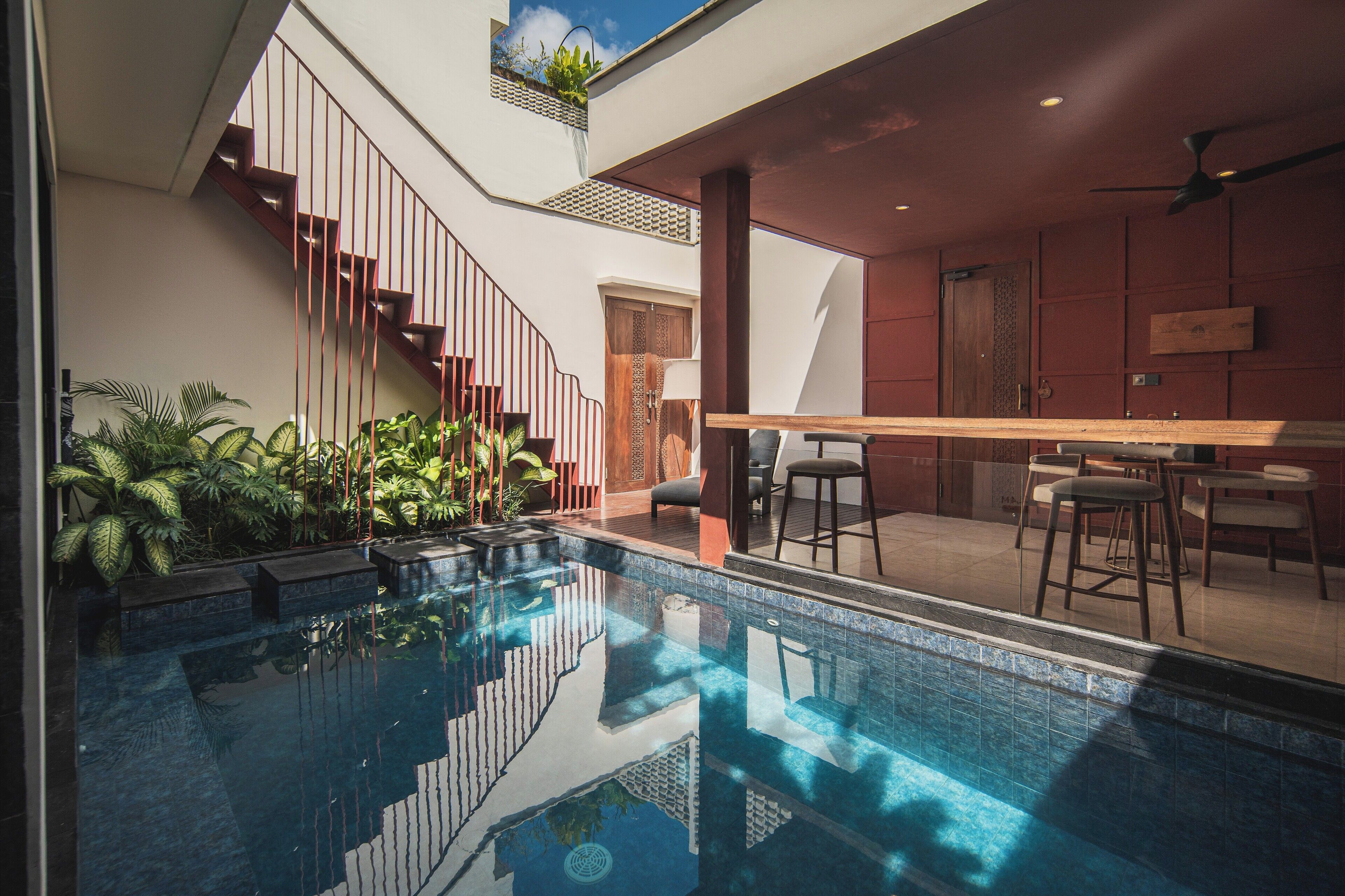 Un espace piscine intérieur moderne, doté d’une paroi en verre transparent, d’un escalier en bois et d’un bar avec des sièges.