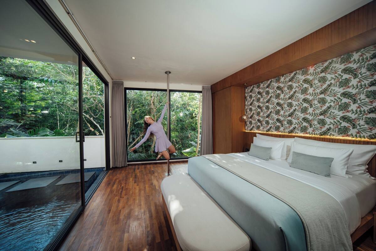 A modern bedroom with a large bed, a wooden headboard, and a view of greenery through a sliding glass door.