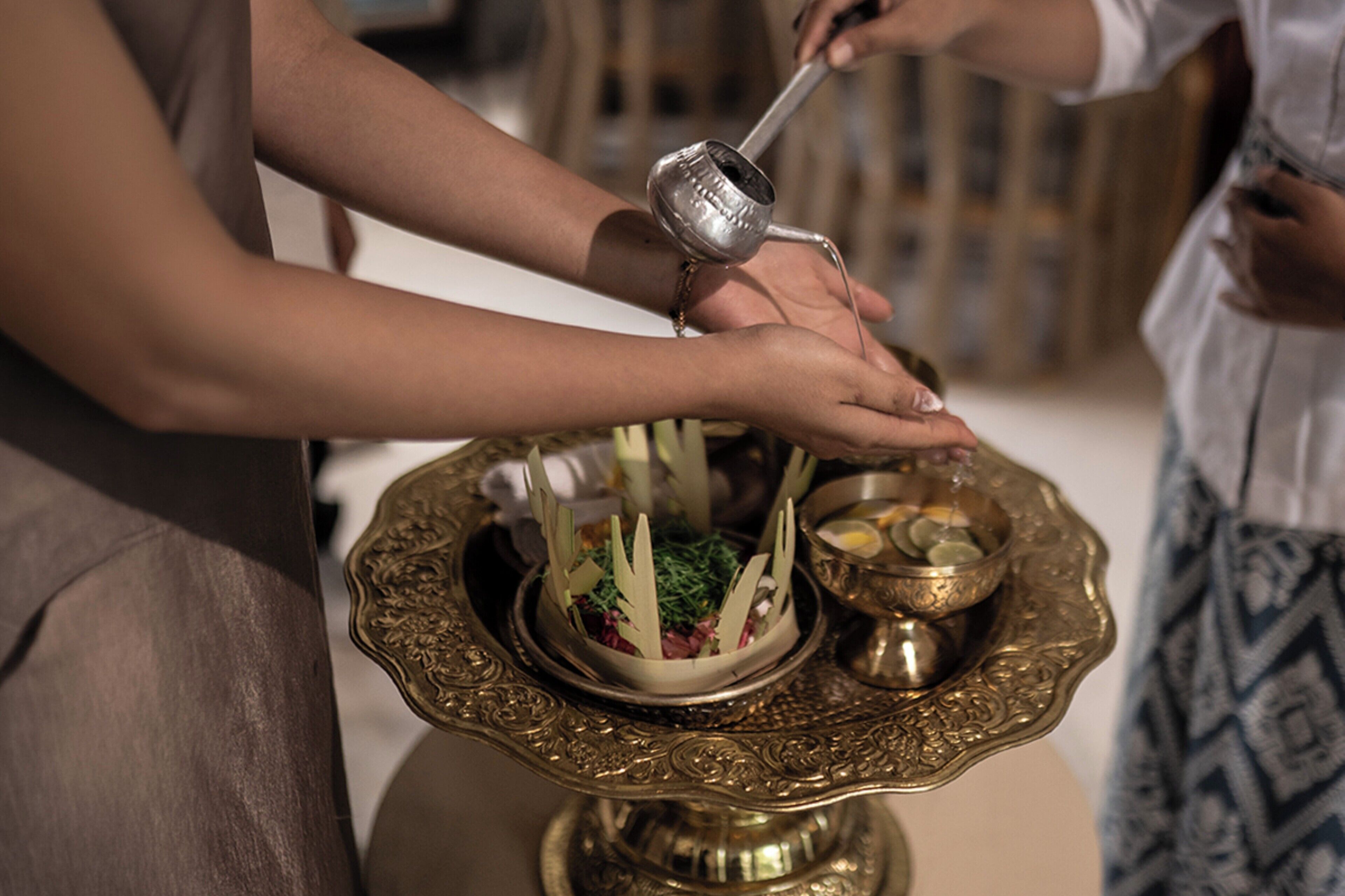 A person pouring a liquid from a metal pitcher into a decorative bowl on a table.