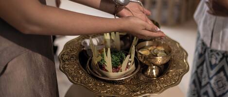 A person pouring a liquid from a metal pitcher into a decorative bowl on a table.