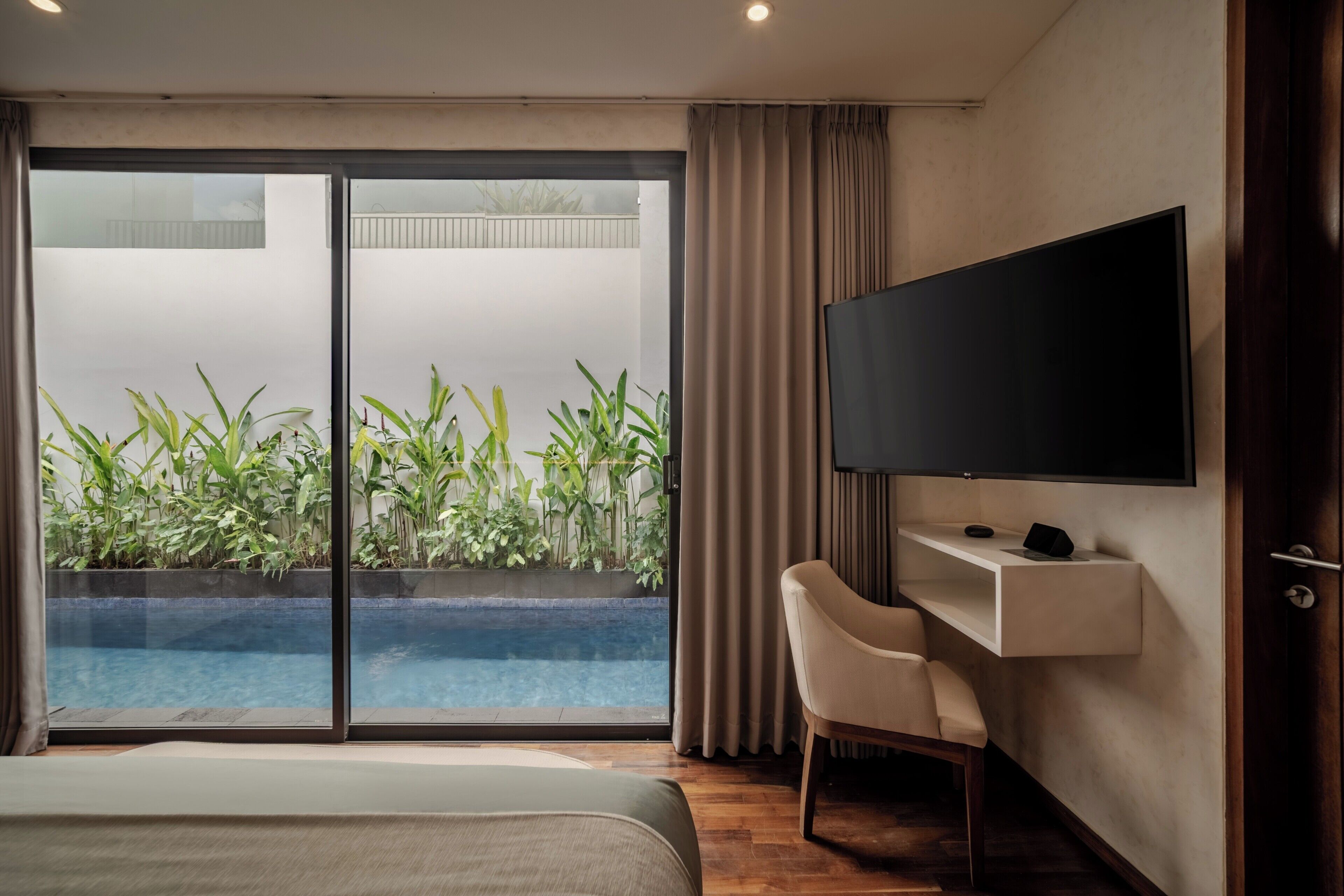 A hotel room with a bed, a desk, a television, and a sliding glass door leading to a pool area with plants.