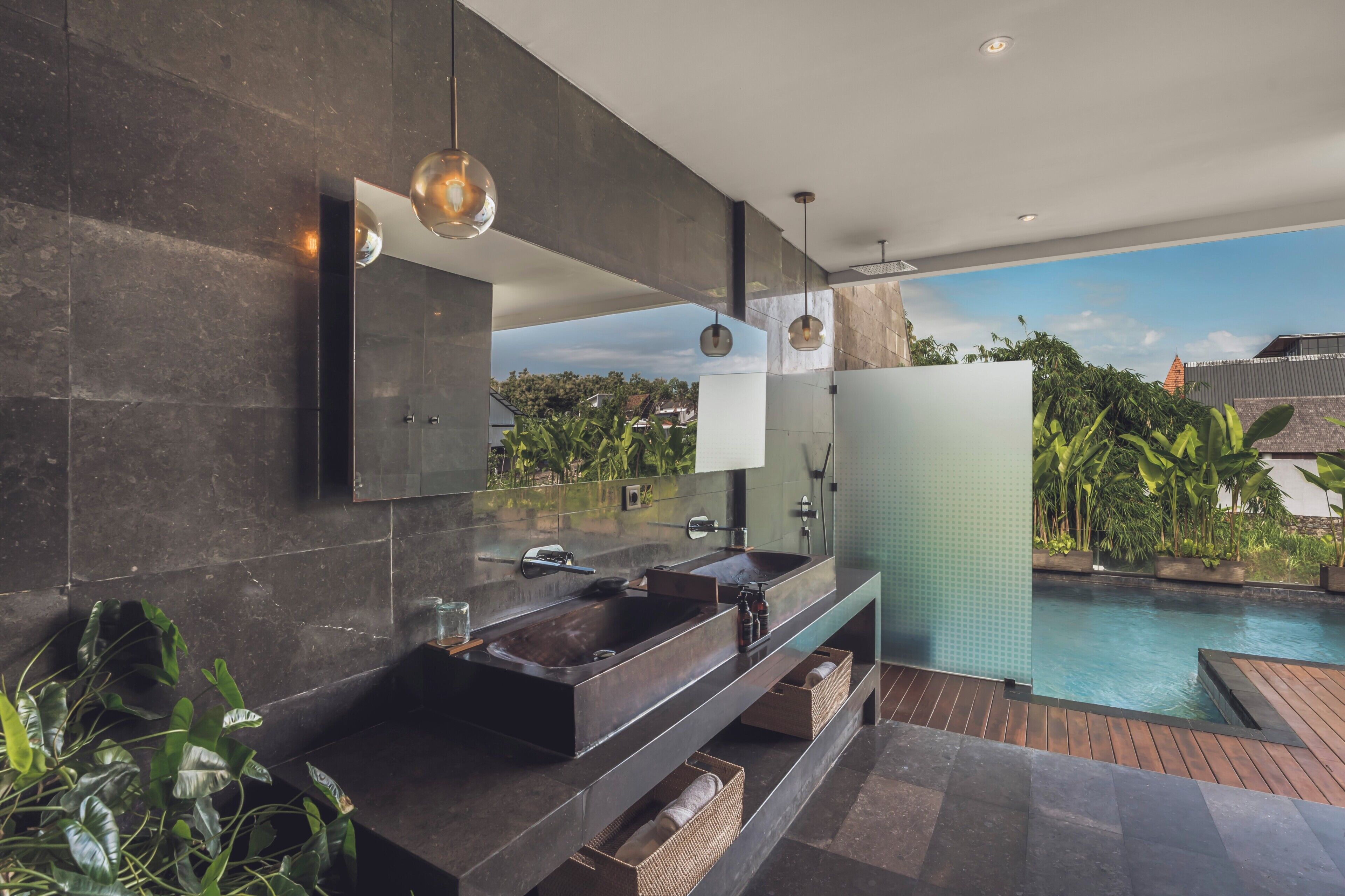 A modern bathroom with a large mirror, dark tiles, a sink, and a view of a swimming pool.