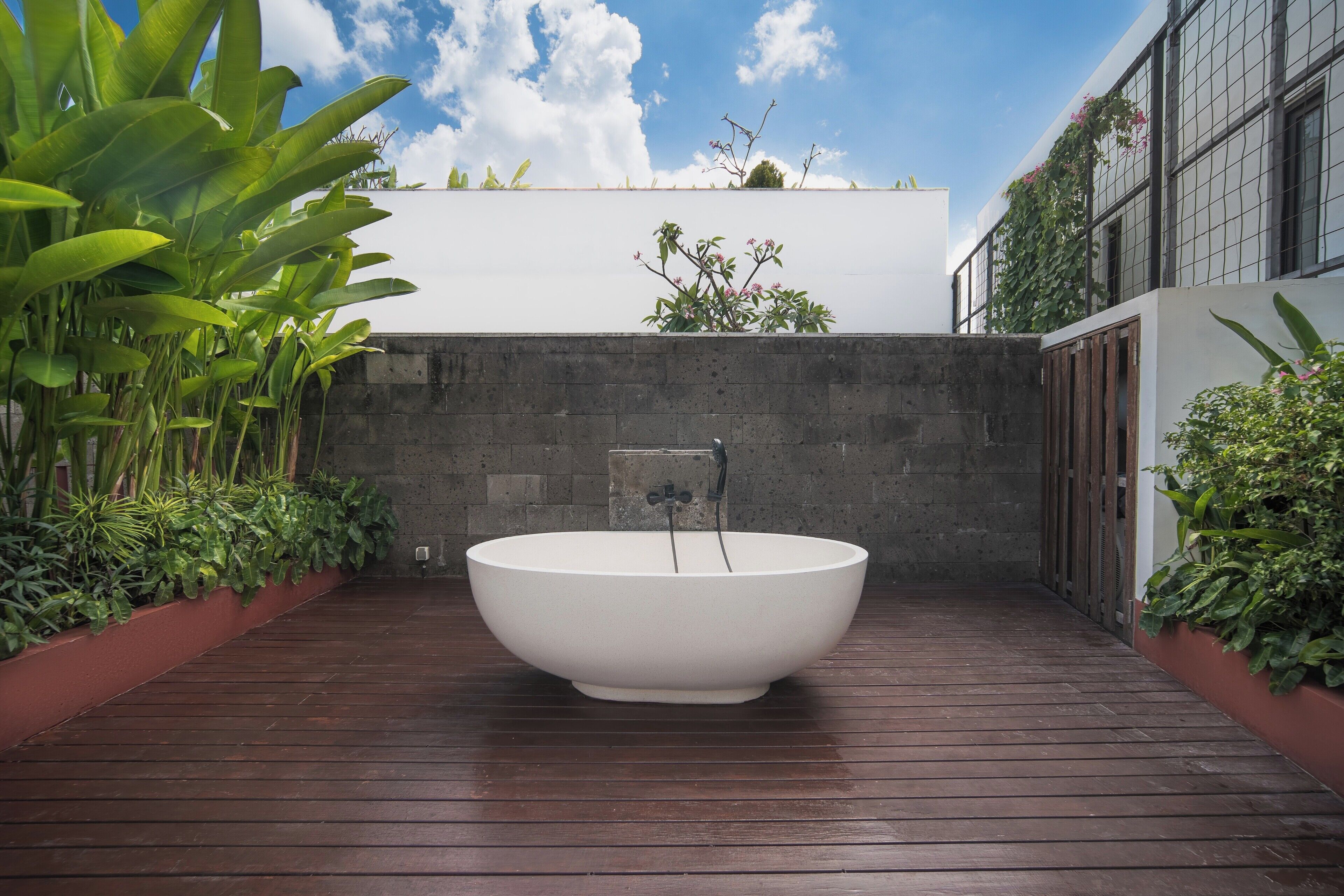 Una bañera blanca grande en un área de terraza de madera, rodeada de follaje y una pared blanca con una ventana.