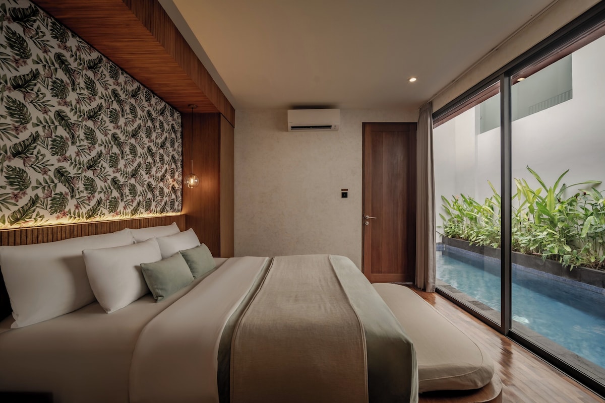 A bedroom with a bed, a wooden headboard, a wall with a leaf pattern, a sliding door to a balcony, and a view of a swimming pool.