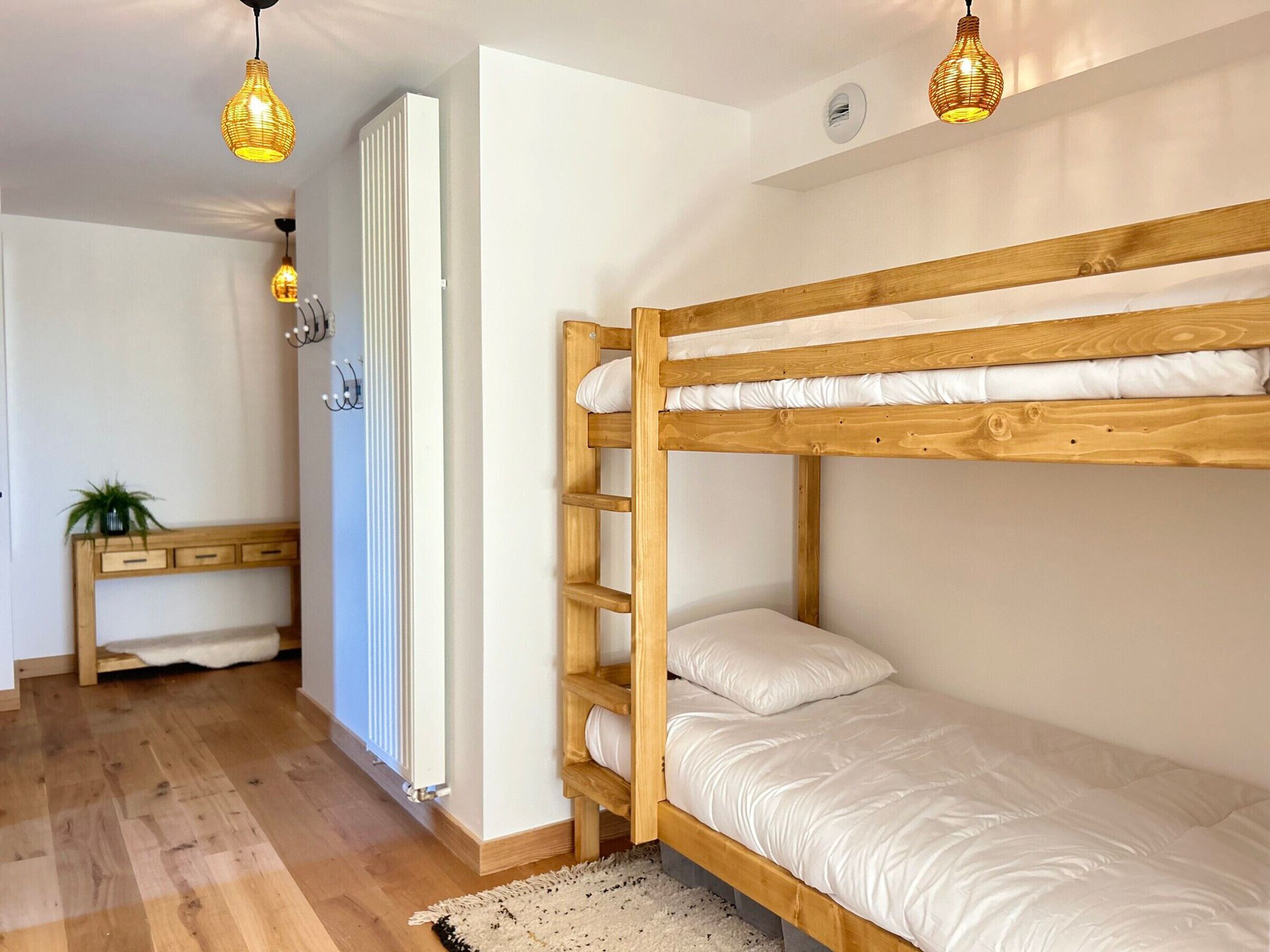 A bunk bed with a wooden frame and white bedding, a small wooden desk with drawers, a radiator, and two hanging pendant lights.