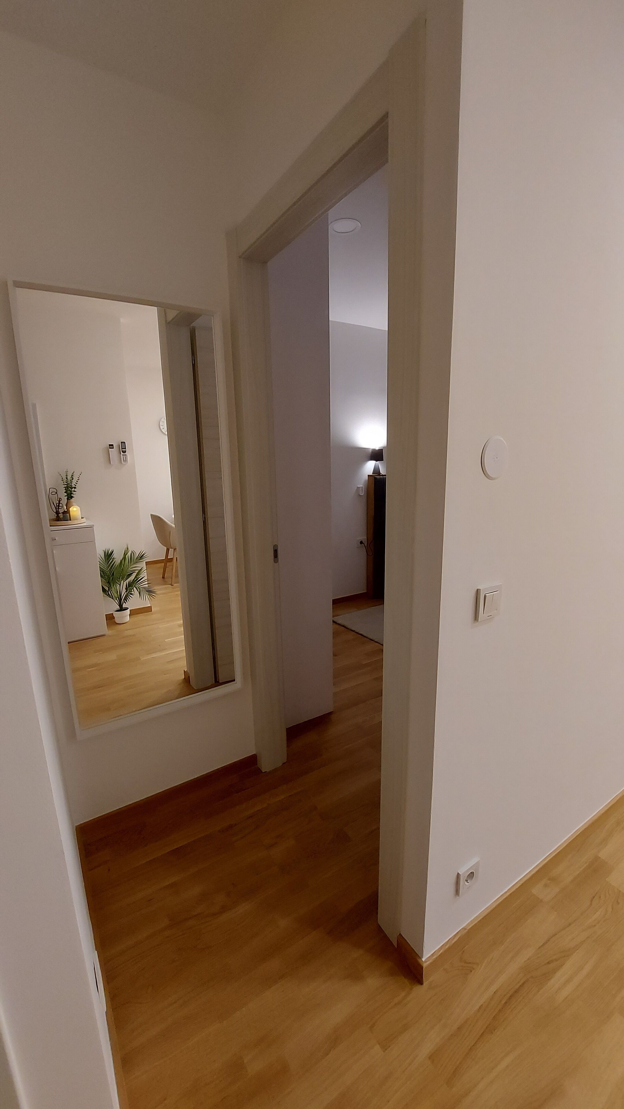 Un pasillo con piso de madera y paredes blancas. Se ve una puerta que da a otra habitación con una lámpara.