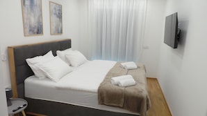 Una cama bien hecha con sábanas blancas y un plaid marrón, una mesita de noche con un control remoto, y un televisor montado en la pared.
