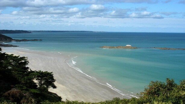 Un paysage côtier avec une plage de sable, des eaux turquoise et limpides, et une petite île rocheuse.