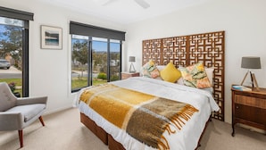 A bedroom with a large bed, a ceiling fan, a window with a view, a chair, and a nightstand with a lamp.