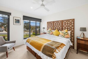 A bedroom with a large bed, a ceiling fan, a window with a view, a chair, and a nightstand with a lamp.