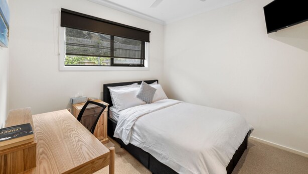 A bedroom with a bed, a desk, a chair, a window with blinds, a ceiling fan, and a mounted TV. - Porta Rossa (Anglesea)