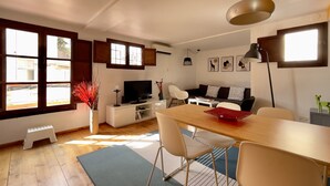 A modern living room with a wooden dining table, white chairs, a TV on a white cabinet, and a sofa with patterned cushions. - Cozy attic penthouse in center of BCN. Sun, views, quiet... close to main spots. (Barcelona)