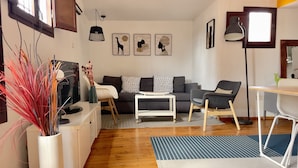 A modern living room with a sofa, armchairs, a coffee table, and a TV. There are framed artworks on the wall, a floor lamp, and a vase with red plants. - Cozy attic penthouse in center of BCN. Sun, views, quiet... close to main spots. (Barcelona)
