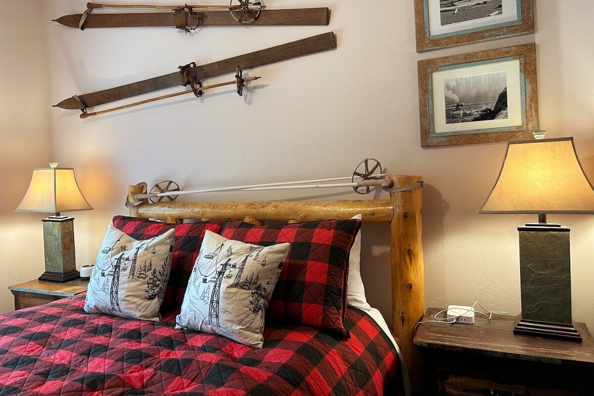 A bed with a red and black quilt, two pillows with industrial designs, a wooden headboard, and a nightstand with a lamp and a power strip.