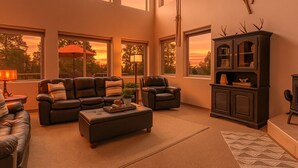 Un salon spacieux comprenant un grand canapé d’angle, deux fauteuils, une table basse et une cheminée. De nombreuses fenêtres laissent entrer la lumière naturelle et offrent une vue sur l’extérieur.