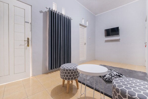 Una habitación con una puerta blanca, una cortina, un televisor montado en la pared y dos otomanas estampadas.
