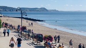 A beach with people walking and sitting on benches, a clear sky, and a calm sea. - Seaward (Lyme Regis)