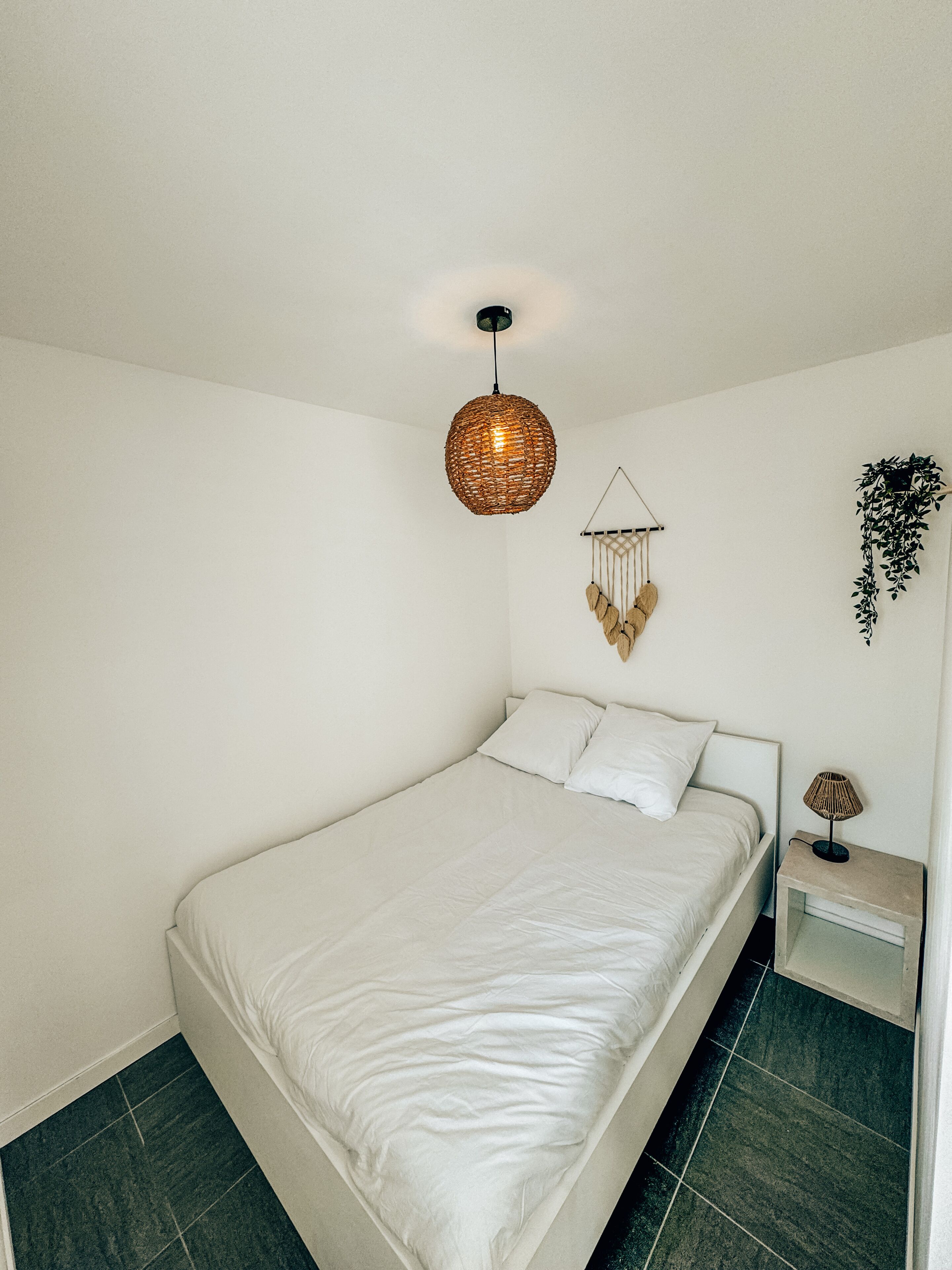 A minimalist bedroom with a single bed, a hanging light fixture, a wall-mounted plant, and a small bedside table with a lamp.