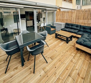 Um pátio externo com uma porta de vidro que dá para uma área de jantar, um deck de madeira e um sofá preto.