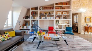 A spacious living room with a high ceiling, a large bookshelf filled with books, a comfortable sofa, a red chair, and a coffee table with a vase of flowers. - Soulful Fusion, by Veeve (Paris)