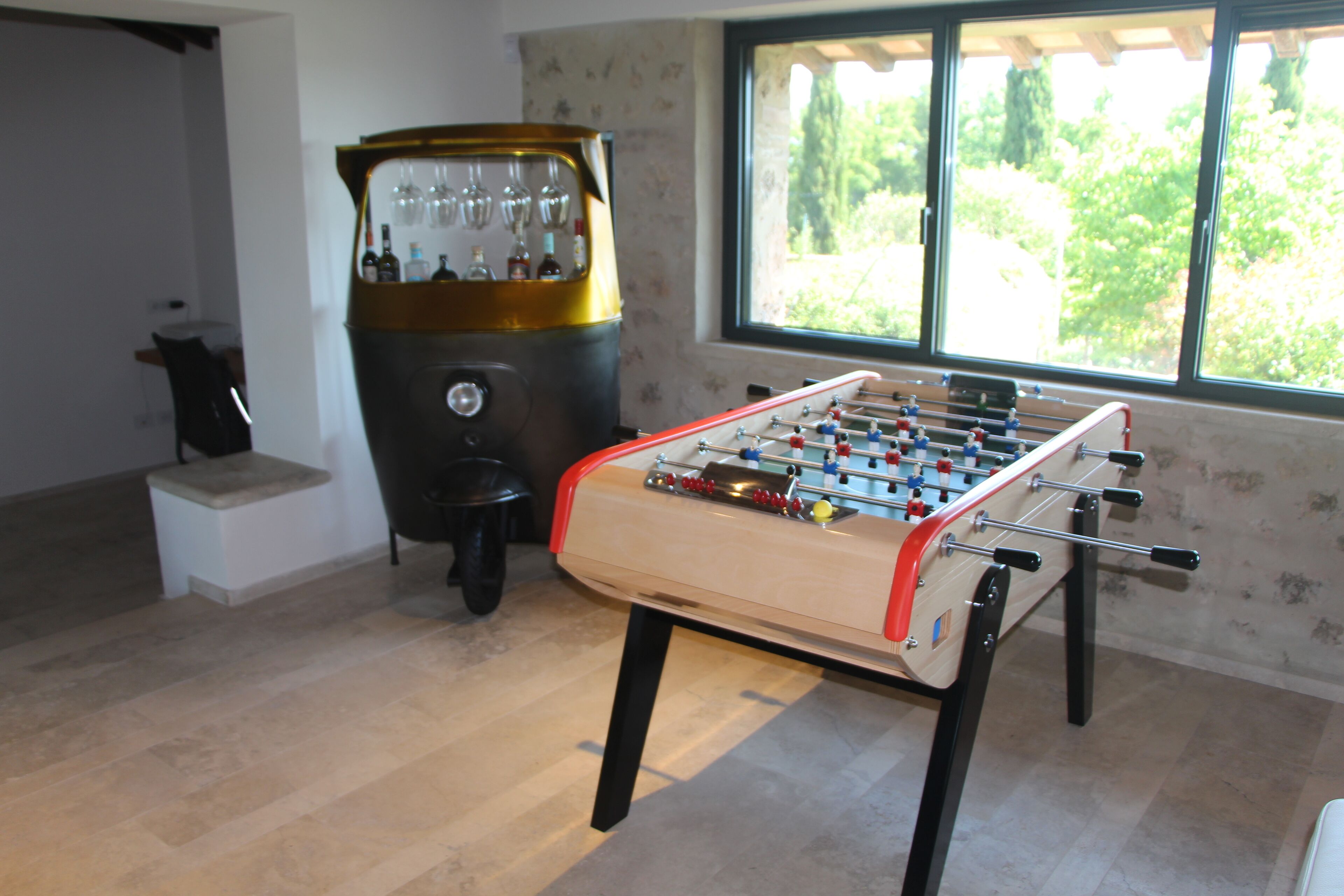 Una mesa de fútbol de mesa en una habitación con ventana, una zona de bar con un mueble dorado y un taburete pequeño.
