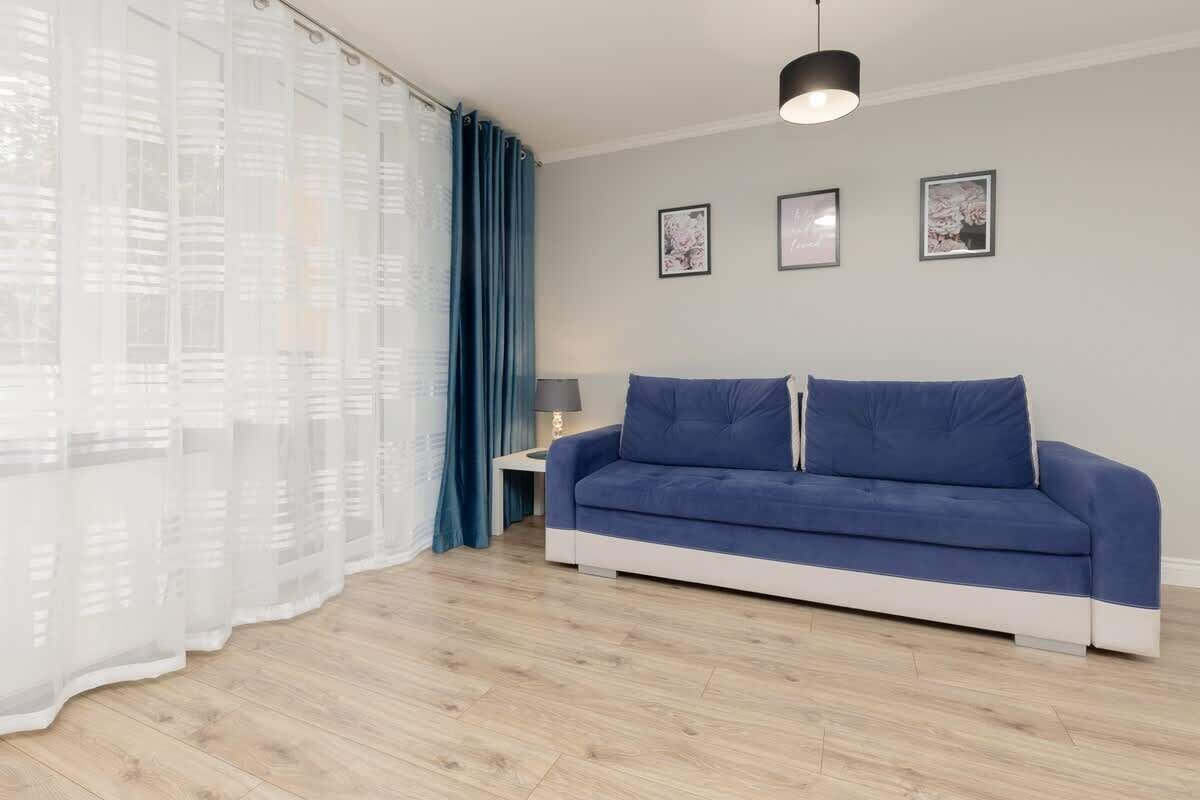 A modern living room with a blue sofa, a side table with a lamp, and a large window with sheer curtains.