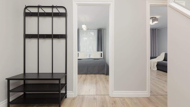 A room with a black metal shelf unit, a bed with a gray bedspread, and a gray sofa in the background. - Sunny Apartment | 2 Bedrooms, Balcony and Air Conditioning (Warszawa)