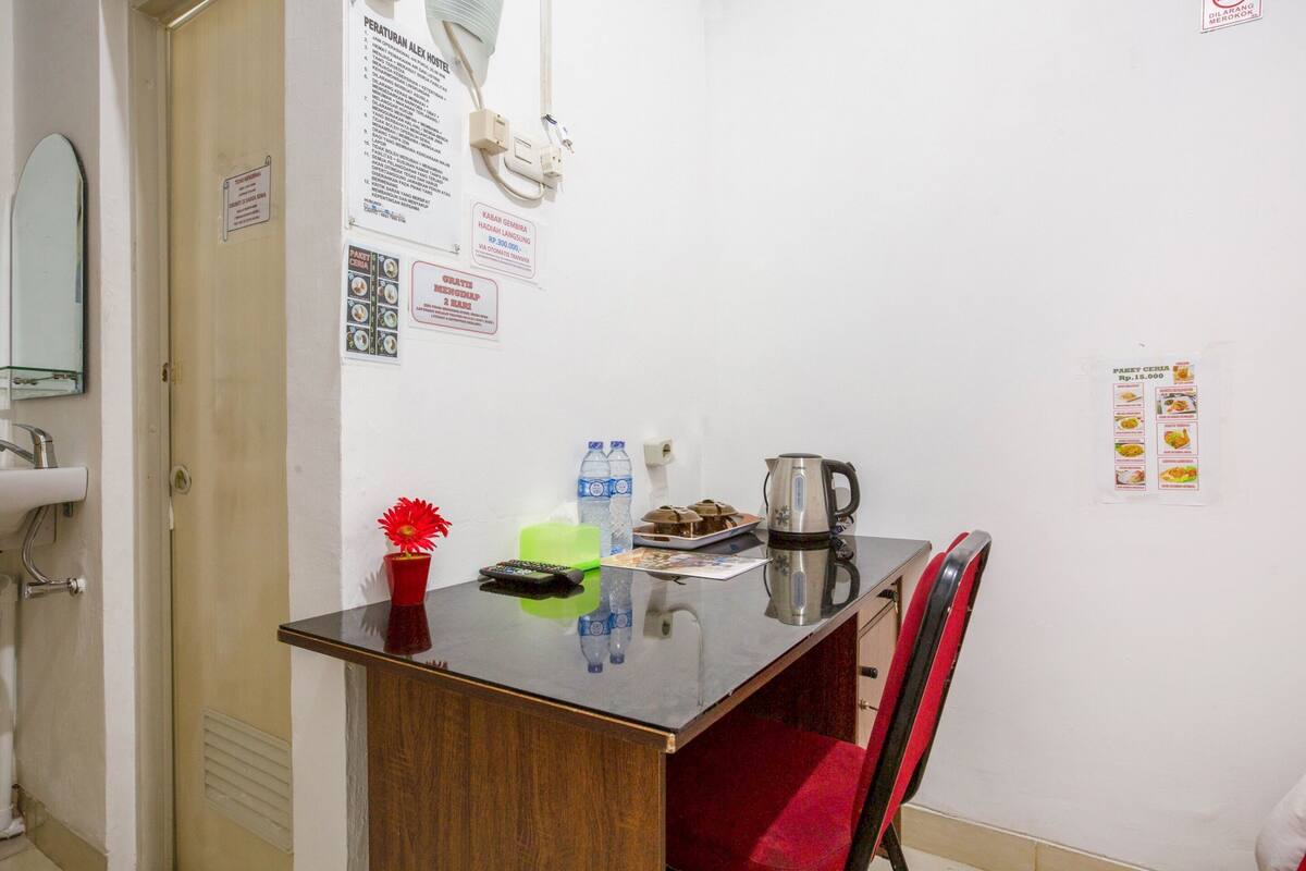 A small room with a wooden desk, a red chair, a sink, and a mirror. There are posters on the wall and a clock.