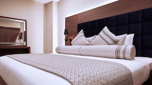 A neatly made bed with a patterned bedspread and pillows, a wooden headboard, and a nightstand with a lamp. - Yogh Hospitality (Paramaribo)