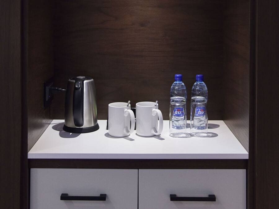 A modern, dark wooden cabinet with a white countertop. On the countertop, there is a silver electric kettle, a white mug, and two water bottles.