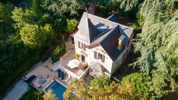 An aerial view of a large house with a swimming pool, surrounded by dense greenery.