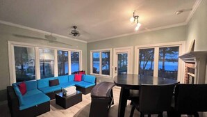 A living room with a blue sofa, a coffee table, a dining table with chairs, and large windows with a view of a body of water. - Modern Lakefront Retreat - 3br 2ba (Sanford)