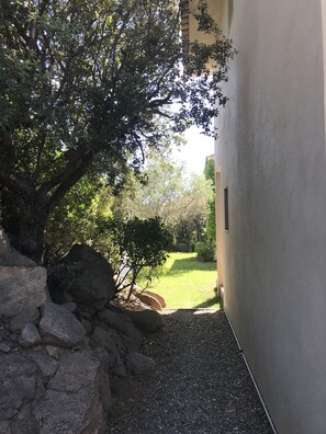 Un chemin pavé mène à un bâtiment dont un côté est bordé d’un mur de pierre et l’autre, d’une zone herbeuse plantée d’arbres.
