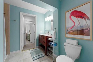A bathroom with a white toilet, a wooden vanity, a mirror, and a framed painting of a flamingo. - Walk to Beach | 3BR beach cottage (Cocoa Beach)