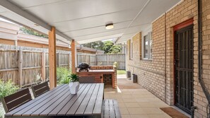 An outdoor patio with a wooden table and benches, a barbecue grill, and a brick wall with a window. - Surfside - Point Lonsdale (Point Lonsdale)